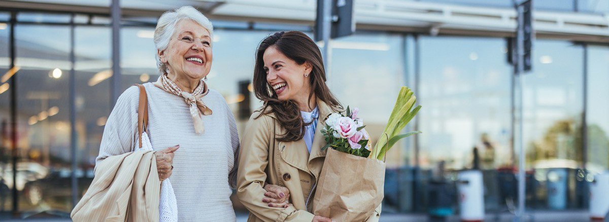 An elderly woman and a young woman smile as they leave the store with their groceries in hand.