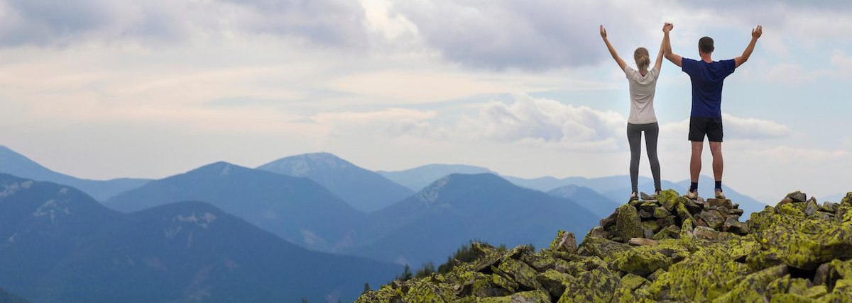 Panorama of couple standing with raised arms on rocky mountain top enjoying summer mountain view