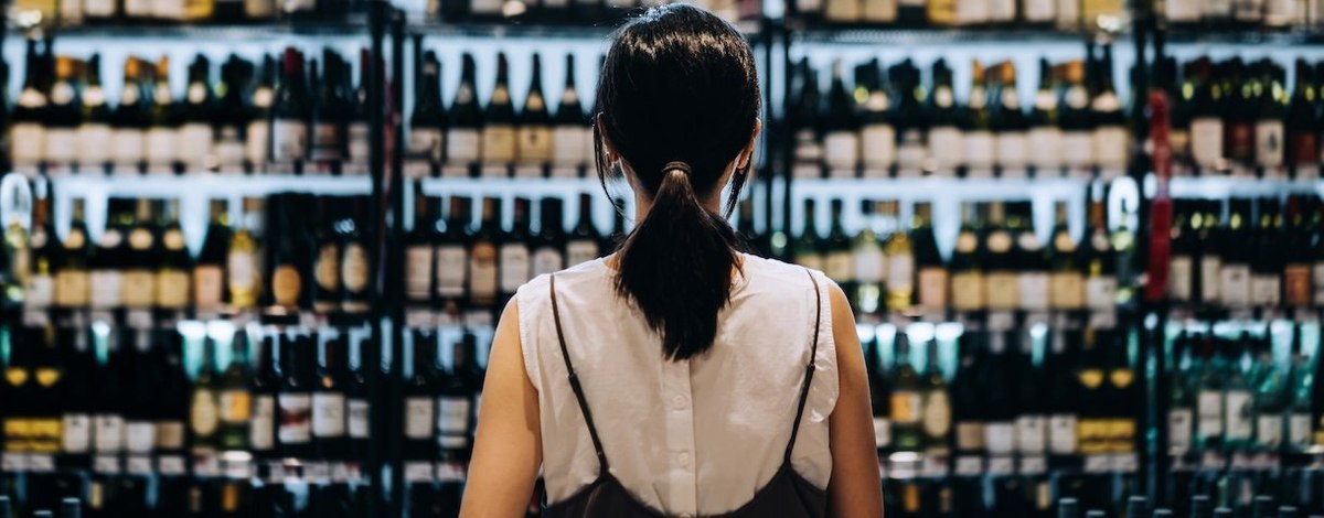 Rear view of young woman grocery shopping for wines in a supermarket. She is standing in front of the liquor aisle and have no idea which wine to choose from