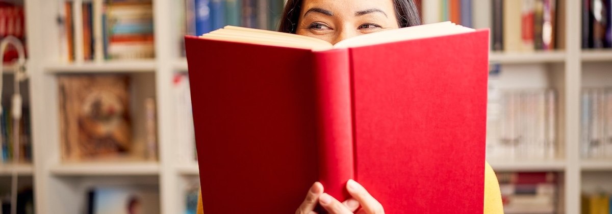 Smiling young woman reading book standing against bookshelf at home