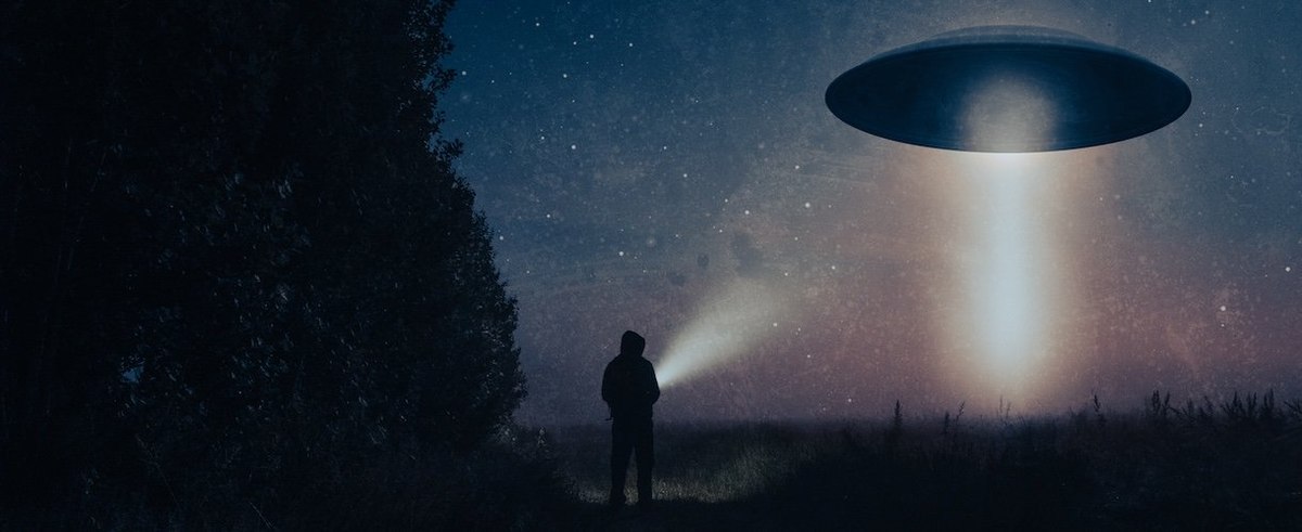 A science fiction concept of a man with a torch looking at an alien UFO. Floating above a field on a spooky foggy night in the countryside.