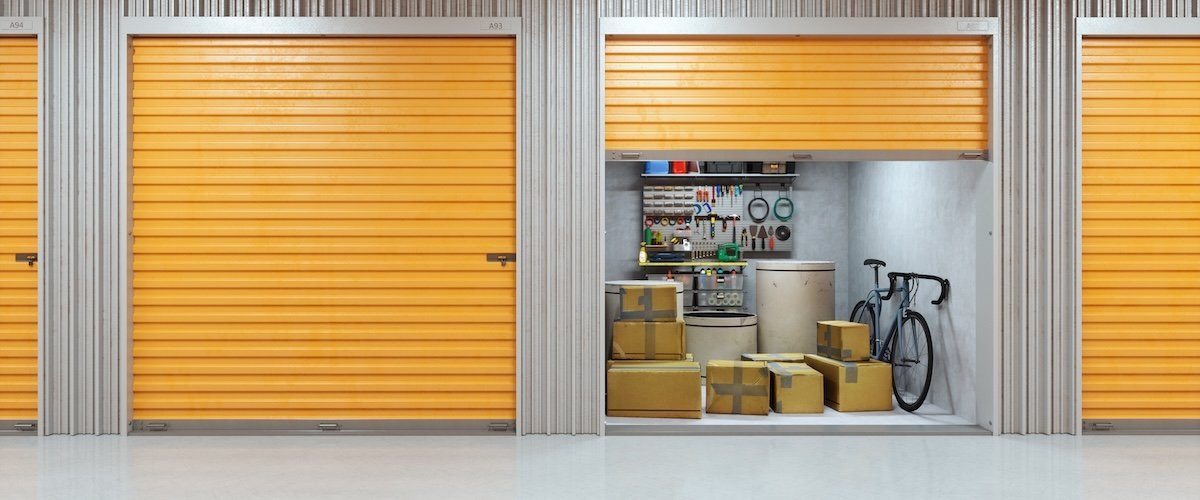 An organized self-storage facility featuring orange storage doors. Inside the unit a bicycle leaning against the wall, cardboard boxes, and an array of tools displayed.