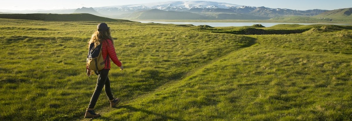 A woman hiking a green grassy trail towards snow covered mountains.